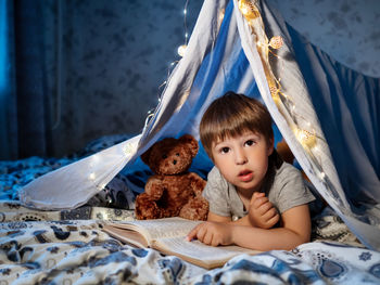 Little boy reads book. toddler plays in tent made of linen sheet on bed. cozy evening with  book.