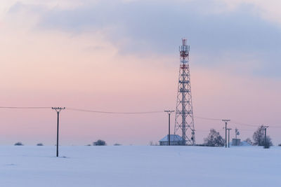 Snow covered electricity pylon against sky during sunset