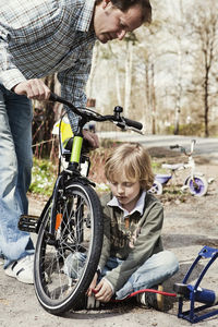 Boy filling bicycle tire with foot pump while father watching him on road