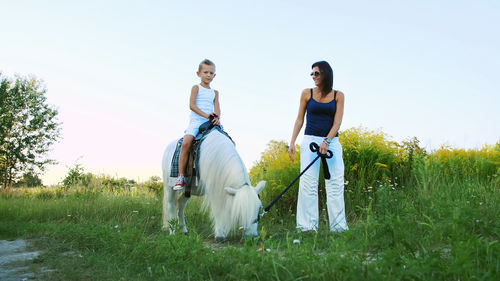 A woman and a boy are walking around the field, son is riding a pony, mother is holding a pony for