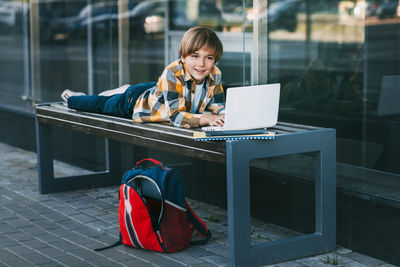 A cheerful boy is lying on a wooden bench and working on a laptop, next to a backpack.