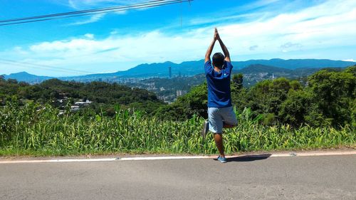 Rear view of man doing tree pose while standing on footpath against sky