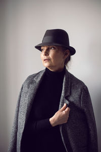 Fashionable older woman in black clothes and a hat stands against a white wall in the office