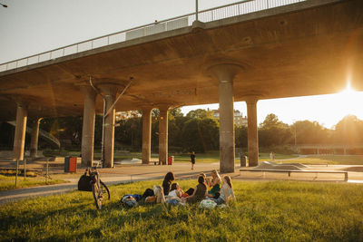 Teenage girls sitting on grass by bridge at park during sunset