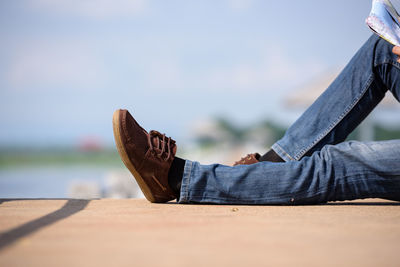 Low section of man wearing shoes while sitting on retaining wall against sky