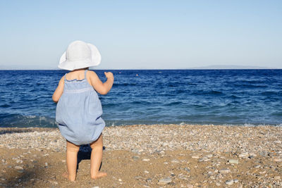 Rear view of baby girl, toddler standing at beach against clear sky, looking at sea