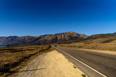 Road leading towards mountains against clear blue sky