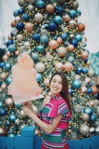 Portrait of smiling young woman standing by christmas tree holding cotton candy outdoors