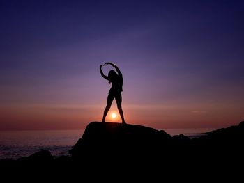 Silhouette woman with arms raised standing on rock by sea during sunset