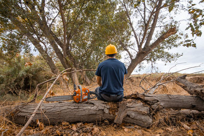 Rear view of man sitting on tree trunk with chainsaw in forest