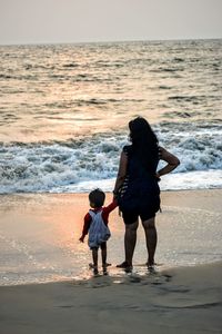 Rear view of mother and girl standing at beach during sunset