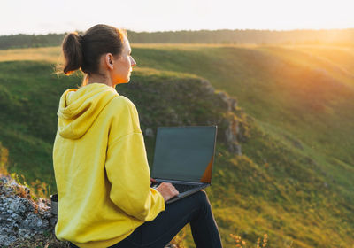 Young woman traveler freelancer in yellow hoodie with opened laptop on beautiful view background