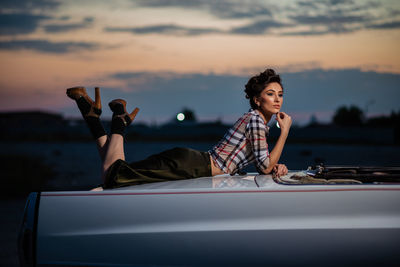 Woman sitting on car against sky during sunset