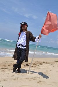 Full length of man in pirate costume holding flag at beach against sky