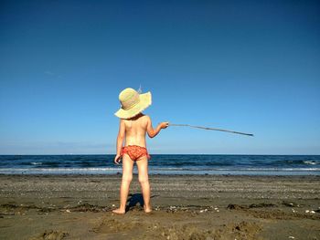 Rear view of girl with stick standing at beach against clear sky