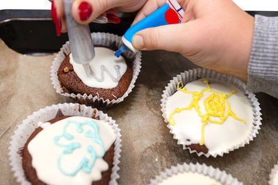 A child squeezes colored frosting from a tube onto chocolate brown cupcakes covered white frosting.