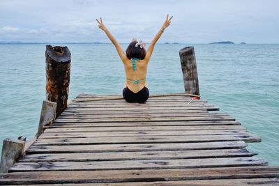 Rear view of woman gesturing peace sign while sitting on pier over sea