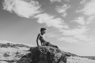 Side view of young woman sitting on rock against sky