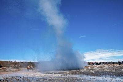 Explorers looking at strokkur geyser eruption in valley against blue sky