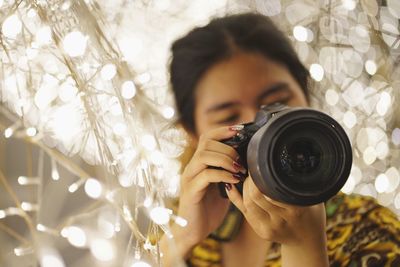 Young woman photographing from camera by illuminated string lights