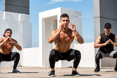 Full length of shirtless men exercising on terrace during sunny day