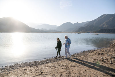 Senior man walking with his granddaughter along the lake shore with mountains.