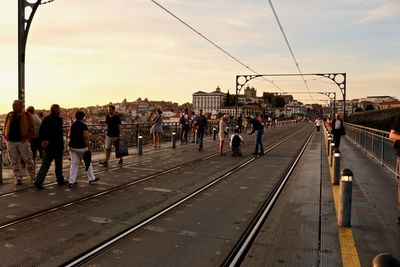 People on railroad tracks in city against sky during sunset