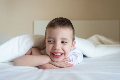 Adorable smiling kid lying down on bed under blanket, looking camera