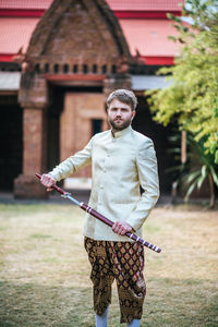 Portrait of young man holding sword while standing on grass against building and sky