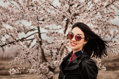Portrait of young woman wearing sunglasses standing against cherry tree