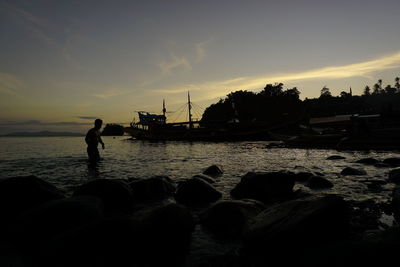 Silhouette man on rock at beach against sky during sunset