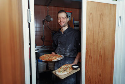 Portrait of young male chef holding pizzas in front of kitchen in restaurant 