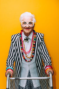 Portrait of stylish senior woman wearing colorful jewelry standing against yellow background