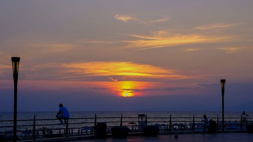 Silhouette people on street by sea against sky during sunset