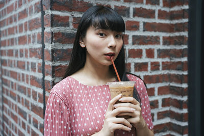 Portrait of young woman drinking cold coffee against brick wall