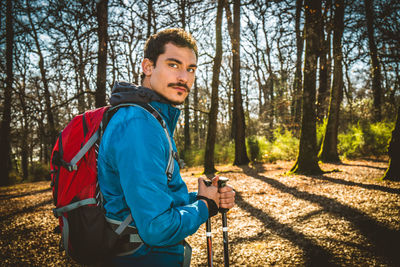 Young man is hiking in the woods. man has a blue jacket, a red backpack and trekking poles.