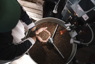 Hands of coffee roaster holding roasted coffee beans near machinery