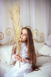 Portrait of girl holding golden feather while sitting on bed