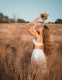 Young woman holding flowers standing on field against sky