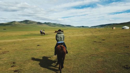 Rear view of man with horse on grassy field against cloudy sky during sunny day