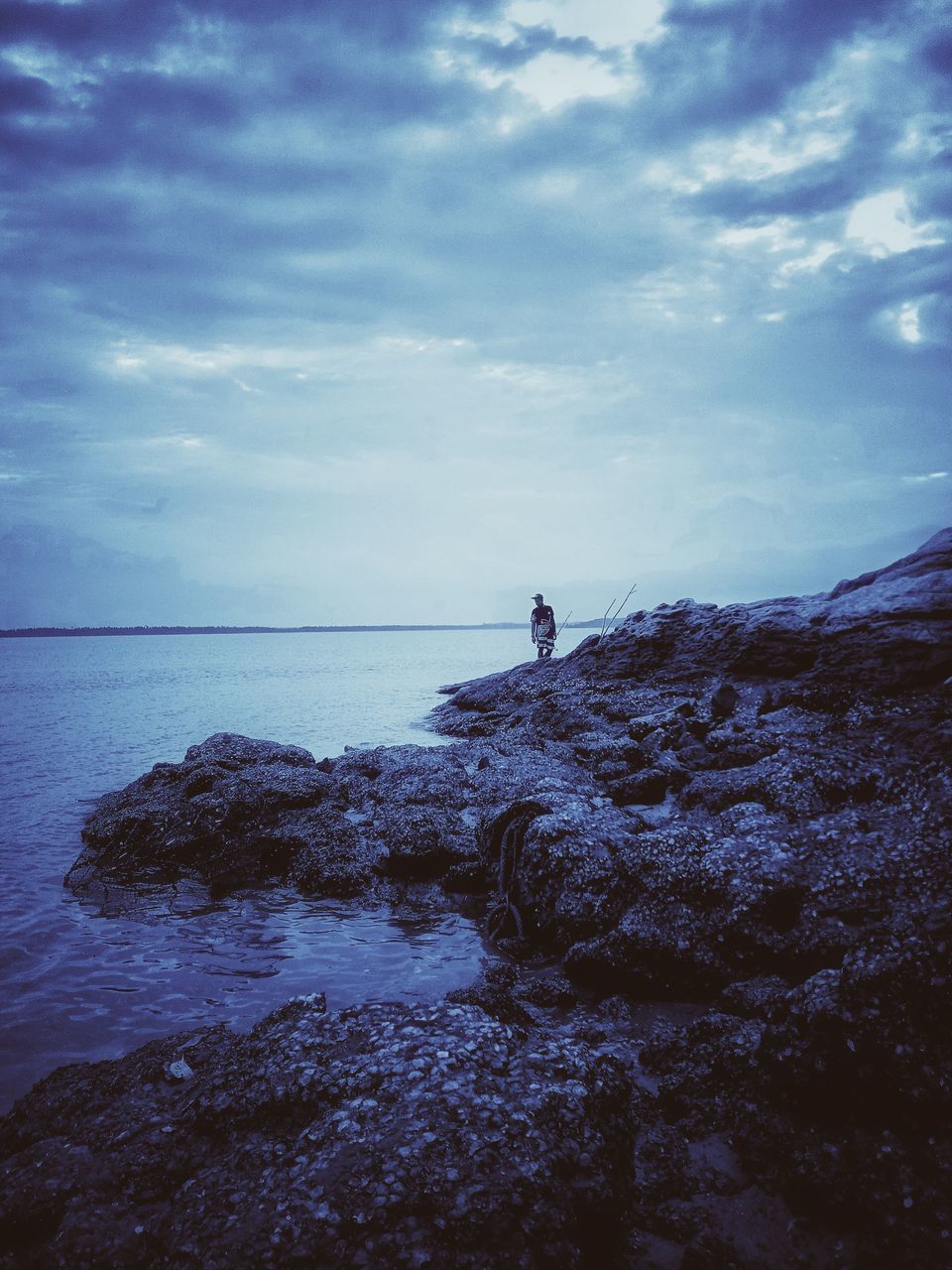 MAN STANDING ON ROCK IN SEA AGAINST SKY