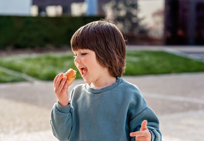 Hungry toddler boy enjoying snack taking a bite while walking outside in park at spring sunny day.