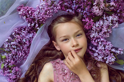 Child girl in a purple floral dress lies on the ground among lilacs on a veil in spring