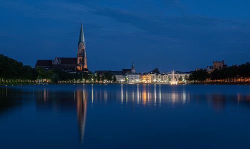 Illuminated buildings by lake against blue sky at dusk