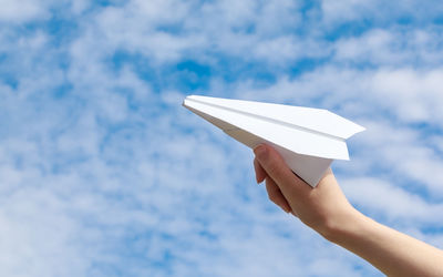 Cropped hand of woman playing with paper airplane against cloudy sky