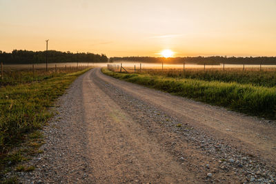Dirt road amidst field against sky during sunset