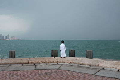 Rear view of man standing on retaining wall by sea against clear sky