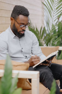 African-american man in an office or coworking makes notes in a notebook and works on a laptop.