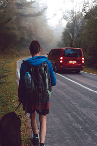 Back view of anonymous female traveler with dog walking along asphalt road near beech forest on foggy autumn day in spain