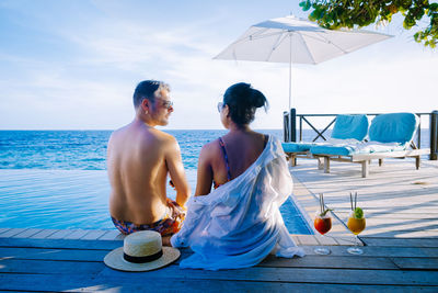 Rear view of couple sitting by swimming pool against sea